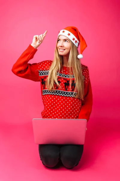 An emotional girl in a red sweater and Santa hat, sitting with a laptop on a red background, depicts that she came up with an idea. — Stock Photo, Image