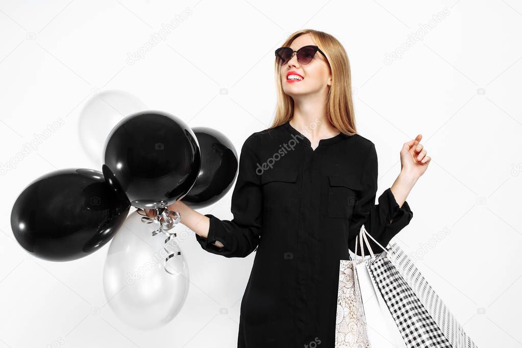 happy woman in sunglasses in black dress and with red lips holding bags and black balls, satisfied girls after shopping, Black Friday on white background