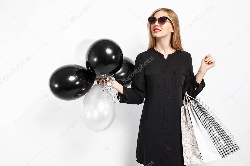 happy woman in sunglasses in black dress and with red lips holding bags and black balls, satisfied girls after shopping, Black Friday on white background