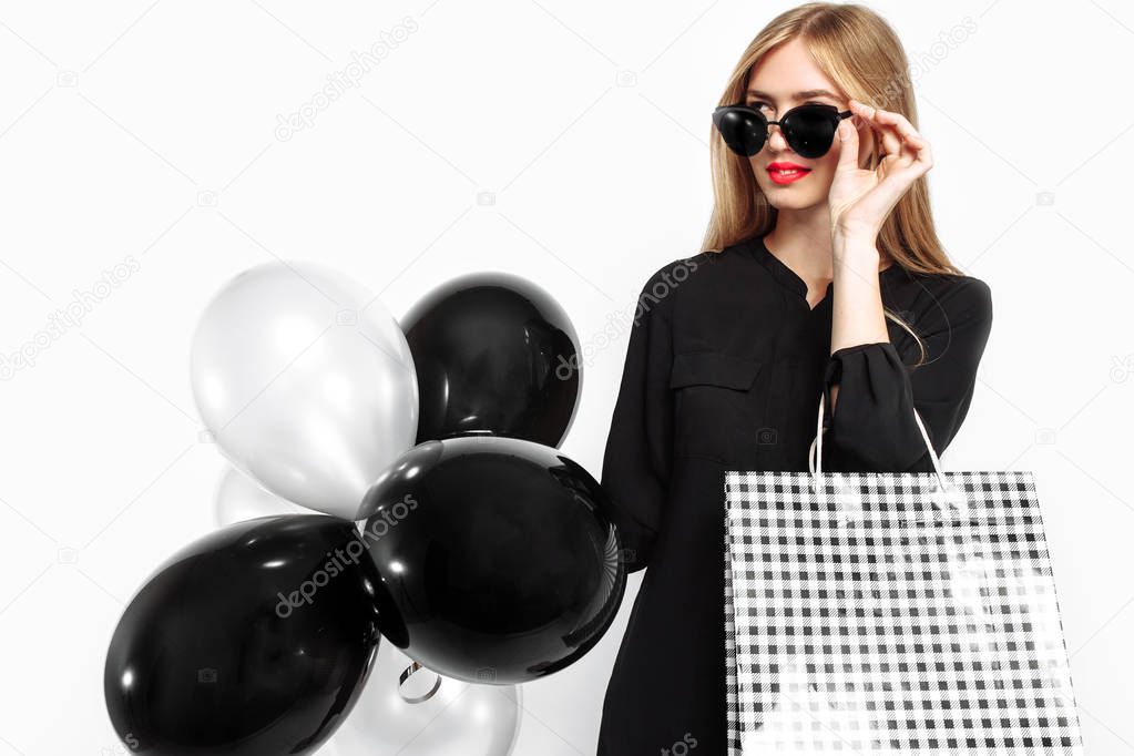 happy woman in sunglasses in black dress and with red lips holding bags and black balls, satisfied girls after shopping, Black Friday on white background