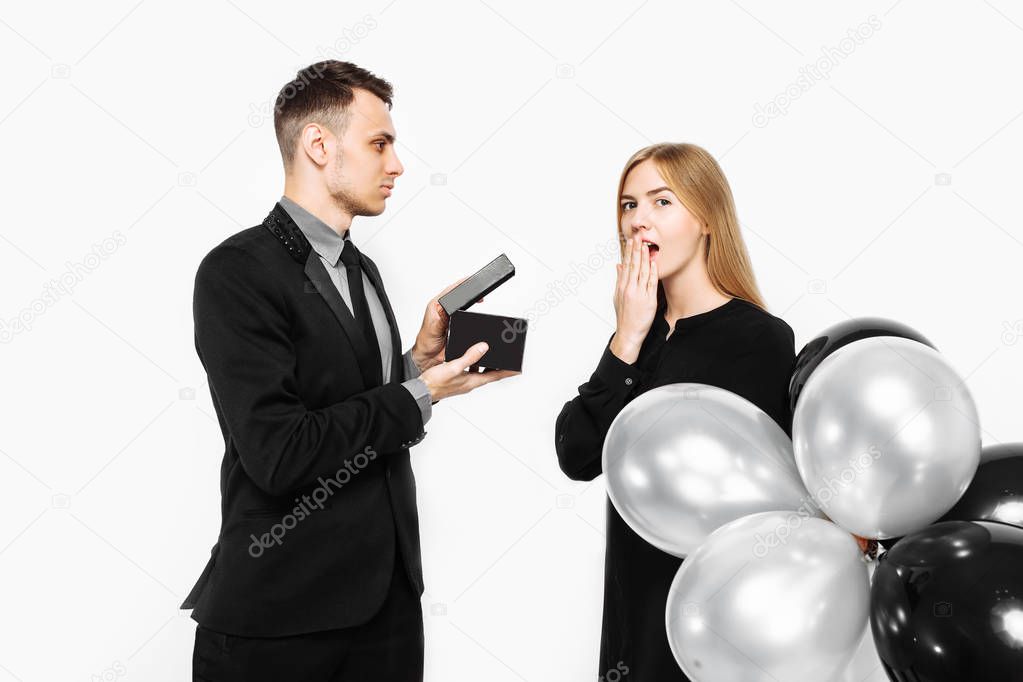 a man in a suit makes a proposal for marriage to a girl who is shocked, a man holds a box with a present in his hands in front of her face, on a white background