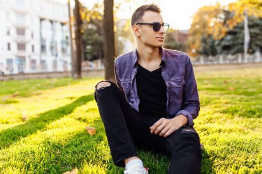 Günbatımında bir park'ın bahçesinde, güneş gözlüğü, rahat kıyafetler giymiş genç bir adam oturuyor.