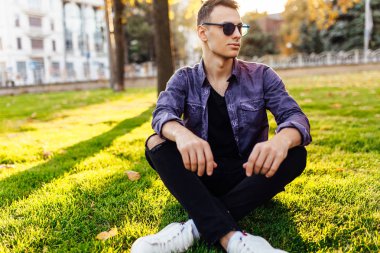 Günbatımında bir park'ın bahçesinde, güneş gözlüğü, rahat kıyafetler giymiş genç bir adam oturuyor.