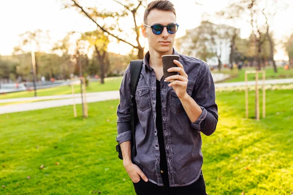 şık giysiler içinde güneş gözlüğü, giyinmiş bir çanta ile çekici bir adam elinde bir bardak kahve tutuyor ve parkta yürüyor,