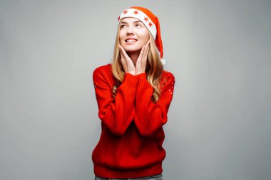 Kırmızı kazak ve ayakta ve gri bir arka plan üzerinde izole yan isteyen bir Noel Baba şapkası giyen bir gülümseyen kadın portresi. Noel