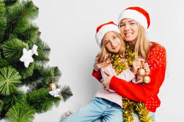 Anne ve kızı Santa Claus şapka, yeni yıl kazak giymiş, Kızı Anne, Noel ağacının yanında oturur hugs. Beyaz bir arka plan üzerinde.