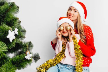 Anne ve kızı Noel Baba şapkaları, yılbaşı kazak giyiyorsun, Noel topları ile oynayarak yılbaşı kutlamak. Beyaz bir arka plan üzerinde.