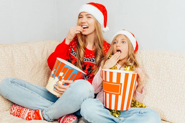 Genç bir anne ve küçük kızı Noel Baba şapkaları, giyinip, film izlemek ve evde kanepede otururken patlamış mısır yemek. Noel kavramı