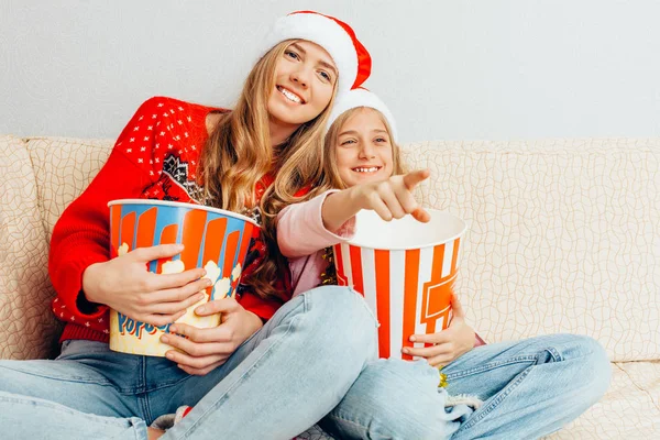 Genç bir anne ve küçük kızı Noel Baba şapkaları, giyinip, film izlemek ve evde kanepede otururken patlamış mısır yemek. Noel kavramı