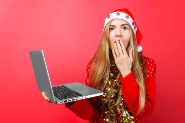 Şok kız kırmızı kazak ve Noel Baba şapkası, elinde, dizüstü bilgisayar ile sürpriz gösteriyor ve ağzını kırmızı bir arka plan üzerinde kapanış kamera bakar