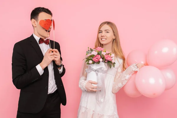 A loving couple, a man holds two paper hearts in his eyes, and a woman holding a bouquet of flowers and balloons in the studio on a pink background