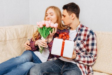 Genç çift aşık adam kadınlar günü kavramı evde kanepede oturan kadın, onu bir buket laleler ve bir hediye vererek kutladı.