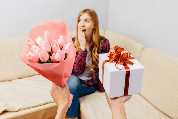 Hands give a bouquet of tulips and a gift to a shocked girl sitting on the couch. Women's day concept