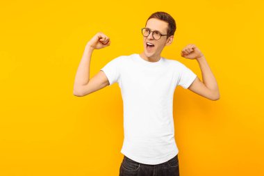 Gözlüklü, beyaz bir T-shirt giymiş mutlu adam kazandı adam bir jest zafer ve başarı, sarı bir arka plan üzerinde gösterir