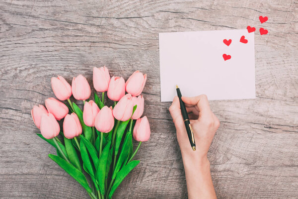 Женские руки пишут на белой бумаге, конверте и красивом букете тонких розовых тюльпанов на деревянном фоне. Весенняя романтика. День святого Валентина
