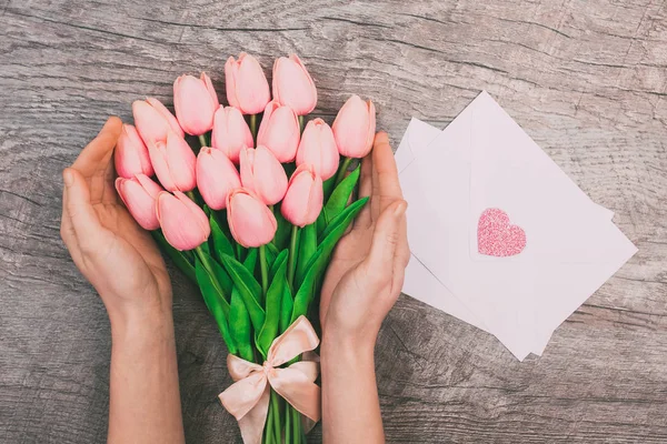 Female hands with a bouquet of pink tulips and clean white envelopes for letters, on a wooden table. View from above. Mother\'s Day or International Women\'s Day.