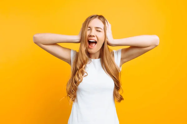 Retrato de uma mulher irritada cobrindo os ouvidos e gritando de um barulho terrível, em um fundo amarelo — Fotografia de Stock