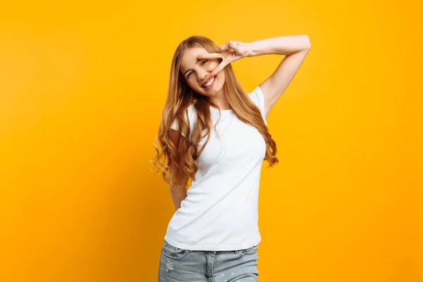 Retrato de uma menina alegre mostrando dois dedos com olhos piscando mostrando língua contra fundo amarelo — Fotografia de Stock