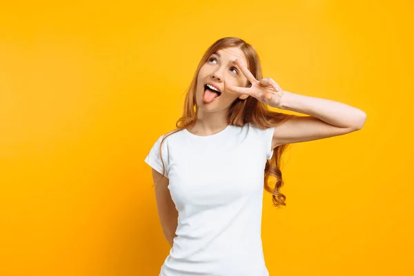 Porträt eines fröhlichen Mädchens, das zwei Finger mit zwinkernden Augen zeigt und die Zunge vor gelbem Hintergrund zeigt — Stockfoto