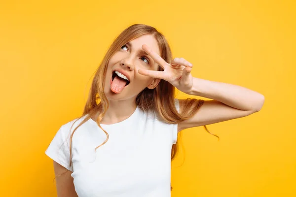 Portret van een vrolijk meisje met twee vingers met winking ogen tonen tong tegen gele achtergrond — Stockfoto