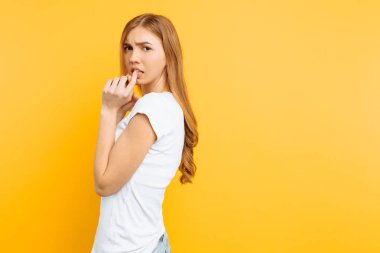 Beyaz tişört, heyecanlı bir kız tırnaklarını ısırır, huzursuz bir yüz ifadesi, sarı bir arka plan üzerinde duruyor,