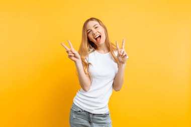 Göz kırpan yanında bir v-sign olarak el hareketi, dil gösterilen ve sarı bir arka plan üzerinde kameraya bakarak bir beyaz T-shirt giyen bir neşeli, eğlenceli kız portresi