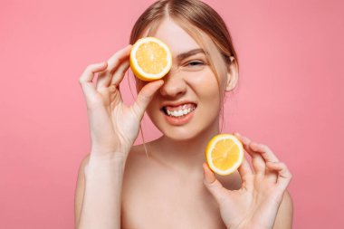 Neşeli bir kadınsı kız, doğal açık cilt, pembe bir arka plan üzerinde izole limon, tek gözüyle kapsayan bir kız iki adet limon, portresi. Cilt bakım kavramı, doğal güzellik