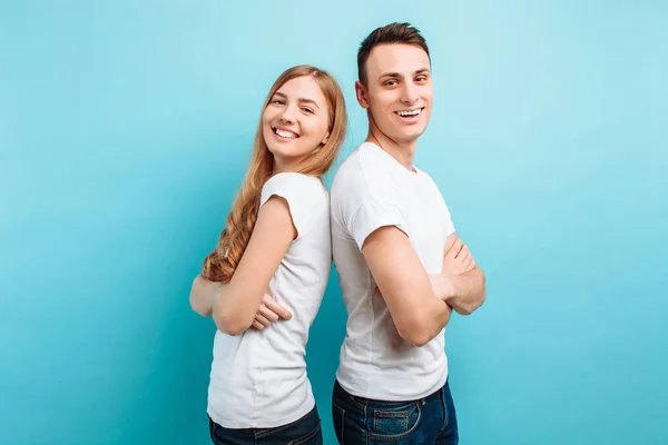 Loving couple, man and woman, dressed in white T-shirts, back-to-back and smiling, against a blue background — Stock Photo, Image