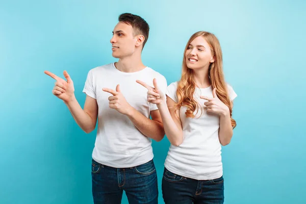 Van een vrolijke jonge paar, man en vrouw wijzen vingers op ruimte voor tekst, geïsoleerd tegen lichte blauwe achtergrond — Stockfoto