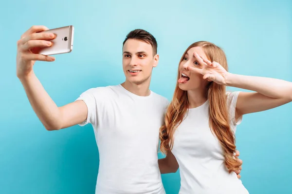Ett lyckligt par män och kvinna tar selfie på en smartphone och ler brett, visar två fingrar över blå bakgrund — Stockfoto