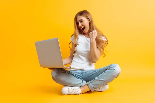 Joven mujer feliz está sentado usando el ordenador portátil y celebrando la victoria y el éxito sobre el fondo amarillo —  Fotos de Stock