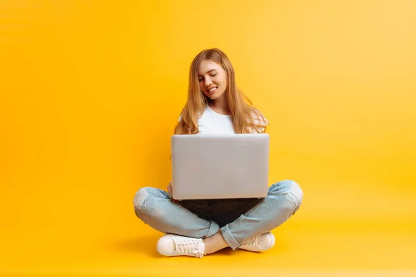 Retrato de una joven sonriente, sentada con las piernas cruzadas usando un portátil, sobre un fondo amarillo —  Fotos de Stock