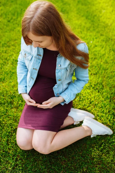 Joyeux jeune femme enceinte assis dans le parc sur la pelouse , — Photo