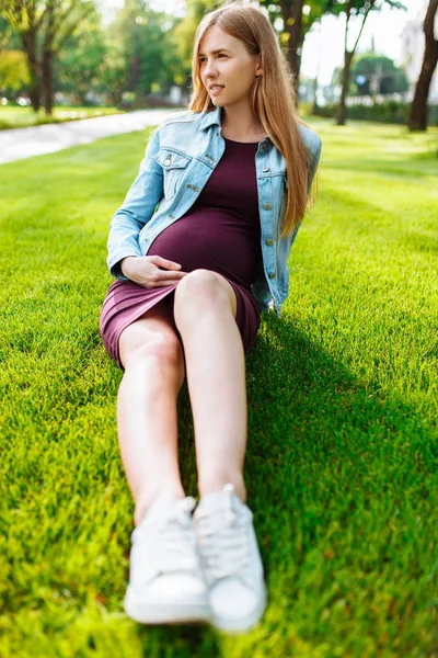 Šťastná Mladá těhotná žena, která sedí v parku na trávníku, — Stock fotografie