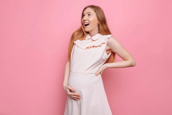 Hermosa mujer embarazada admirando en vestido rosa pone la mano en el vientre sobre fondo rosa — Foto de Stock