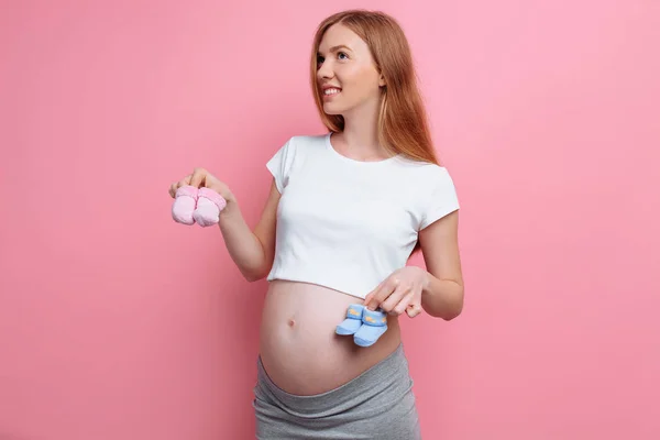 Belle femme enceinte coûteuse tenant des chaussons de bébé bleu et rose, posant en studio sur un fond rose — Photo