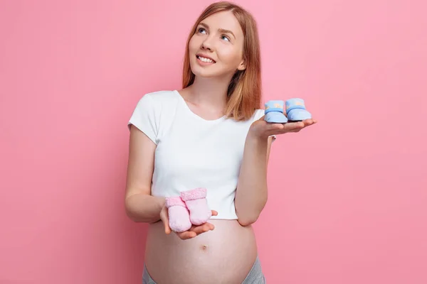 Hermosa mujer embarazada pensativa sosteniendo botines de bebé azul y rosa, posando en el estudio sobre un fondo rosa — Foto de Stock