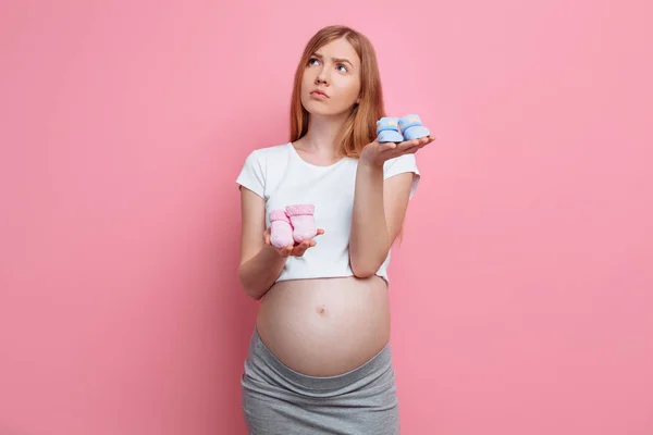 Mooie peinzende zwangere vrouw houdt blauw en roze baby laarsjes, poseren in de studio op een roze achtergrond — Stockfoto