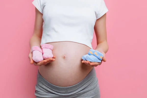 Έγκυος γυναίκα κρατώντας μπλε και ροζ μωρά μποτάκια, ποζάρισμα στο στούντιο σε ροζ φόντο — Φωτογραφία Αρχείου