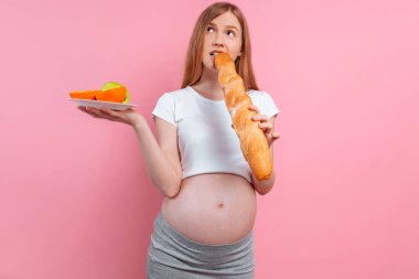 Bir tabak meyve ve pembe arka planda bir somun ekmek ile hamile kadın