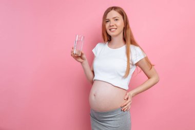 Hamile bir kız, pembe bir arka plan üzerinde, hamile bir karın arka planda bir bardak su tutan