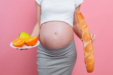 Bir tabak meyve ve pembe arka planda bir somun ekmek ile hamile kadın