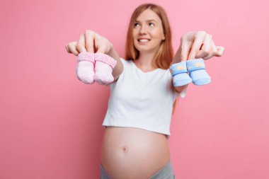 Mavi ve pembe bebek booties tutan güzel dalgın hamile kadın, pembe bir arka plan üzerinde stüdyoda poz