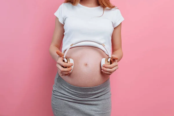 Ελκυστική έγκυος γυναίκα κρατώντας τα ακουστικά στην κοιλιά της πάνω από ένα ροζ φόντο — Φωτογραφία Αρχείου