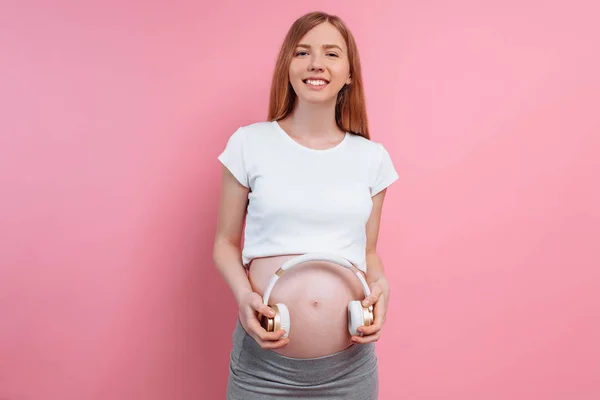 Atrakcyjna kobieta w ciąży trzyma słuchawki na brzuchu na różowym tle — Zdjęcie stockowe