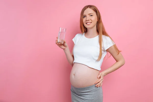 Fille enceinte tenant un verre d'eau sur le fond d'un ventre enceinte, sur un fond rose — Photo