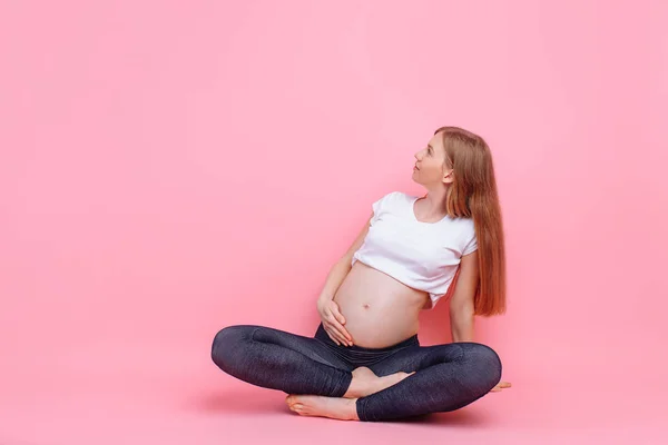 Jong zwanger meisje zitten op een roze achtergrond, op zoek naar de kant van lege ruimte — Stockfoto