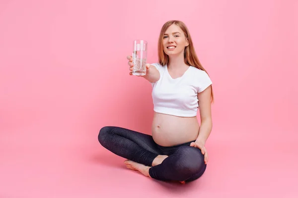 Dziewczyna w ciąży trzymając szklankę wody na tle brzucha w ciąży, na różowym tle — Zdjęcie stockowe