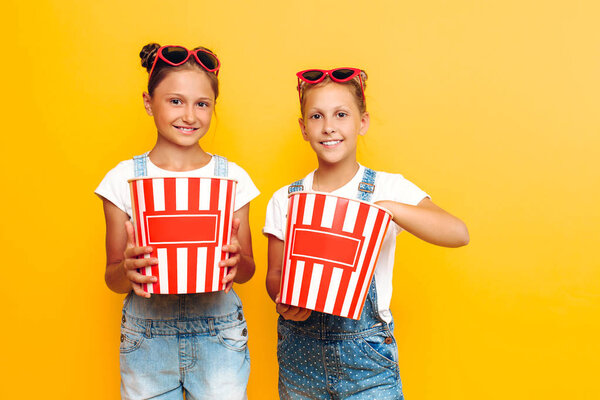 Two teenage girls, girlfriends with popcorn in their hands, posing on a yellow background