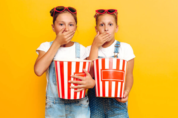 Two teenage girls, stylish girlfriends with popcorn in their hands, watching a horror movie on a yellow background